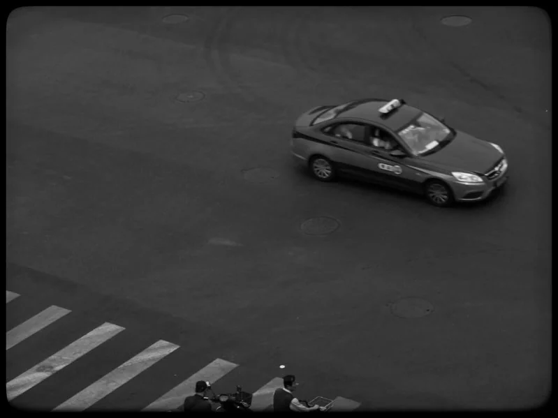 A taxi crossing an empty stretch of road, seen from above, in black and white